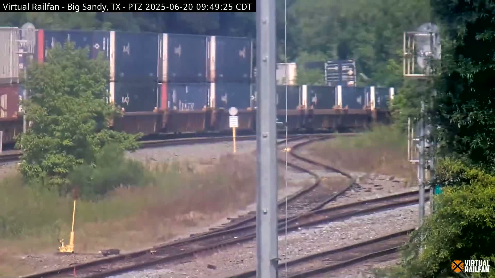 A long freight train carrying stacked shipping containers moves along a curving railway track on a sunny day, passing through a landscape of green vegetation.