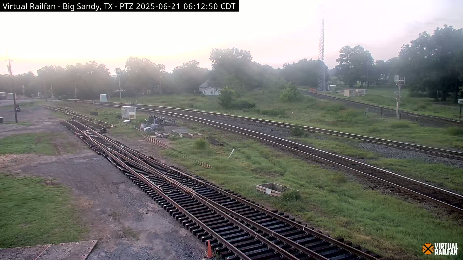 A foggy sunrise illuminates a railway yard with multiple sets of tracks, some under construction, surrounded by grassy fields and trees.