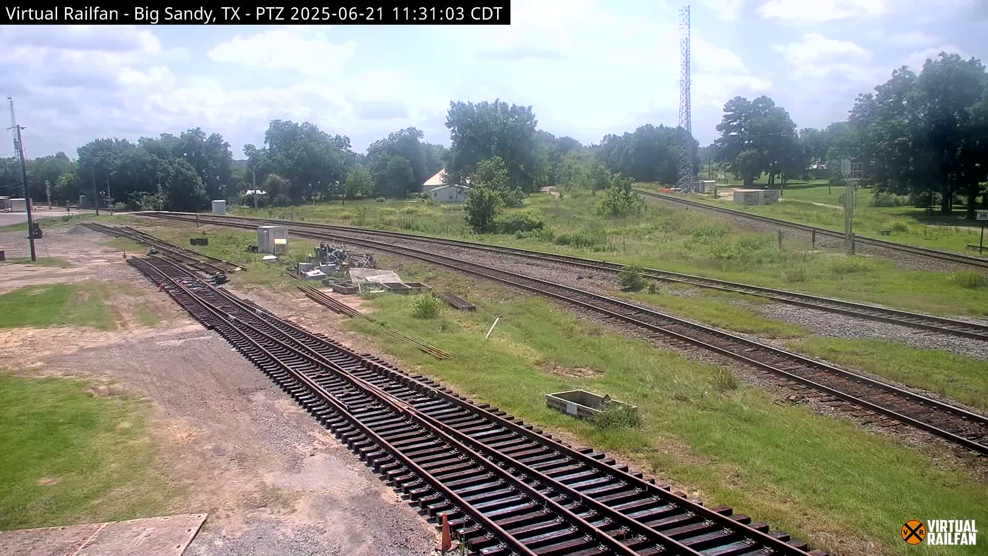 A sunny day shows several sets of railroad tracks winding through a grassy area with trees and a few buildings in the distance.