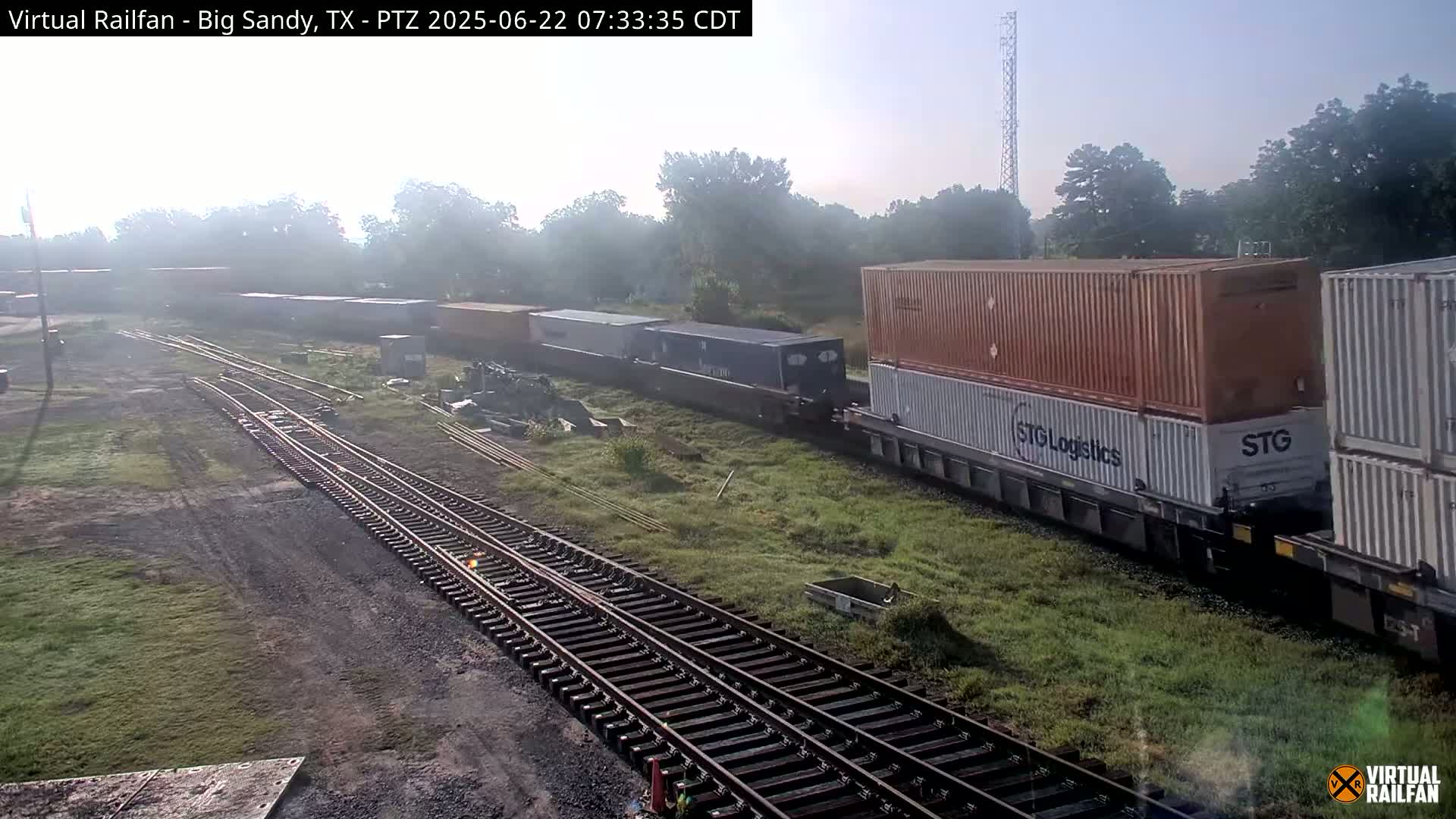 A freight train carrying shipping containers passes alongside railroad tracks on a hazy morning.