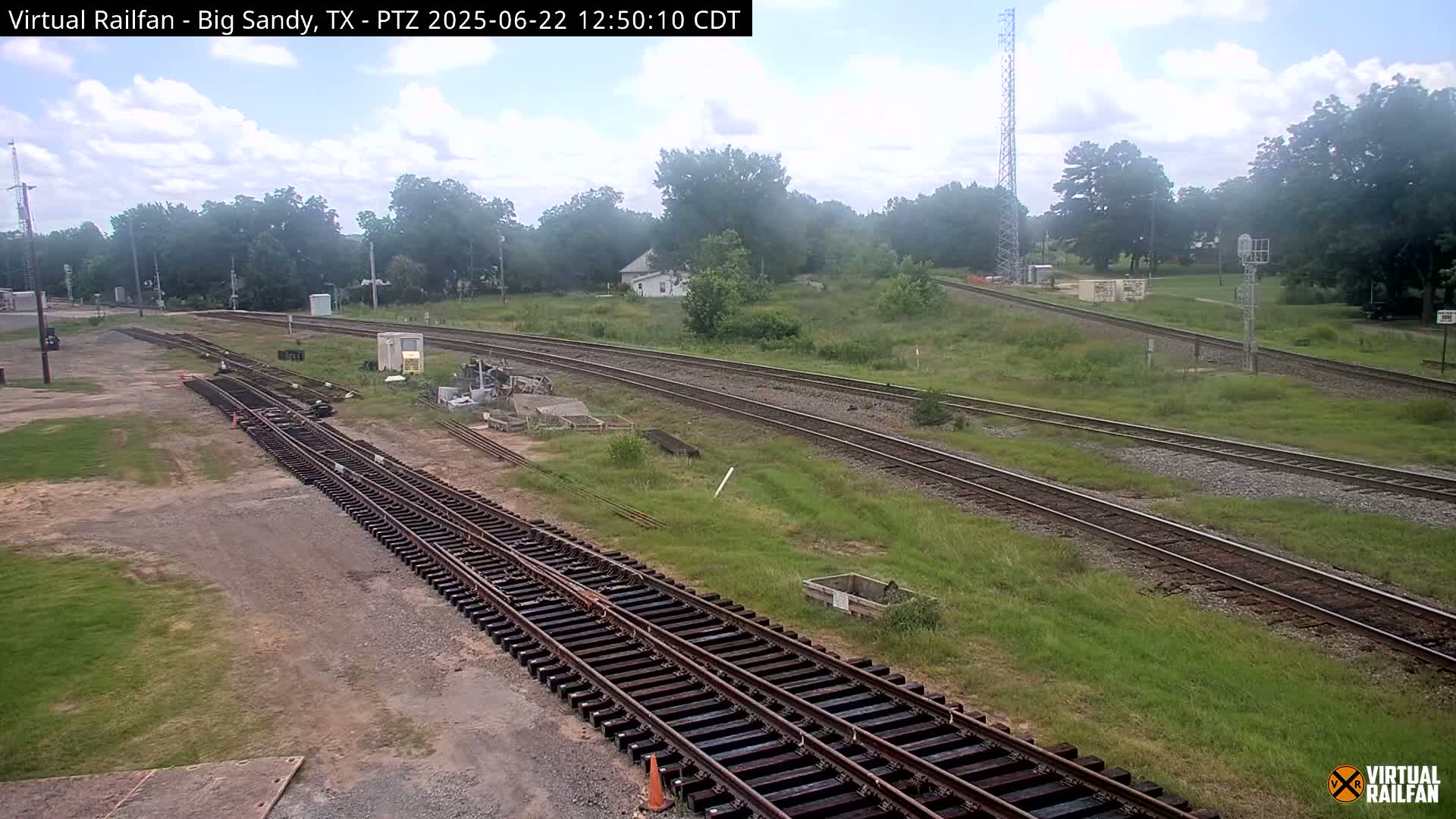 Under a partly cloudy sky, railroad tracks diverge through a grassy area with scattered trees and a small building in the background.