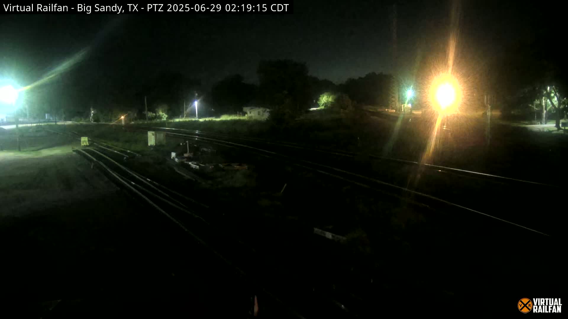At night, several sets of train tracks are visible under artificial lighting, surrounded by dark vegetation.
