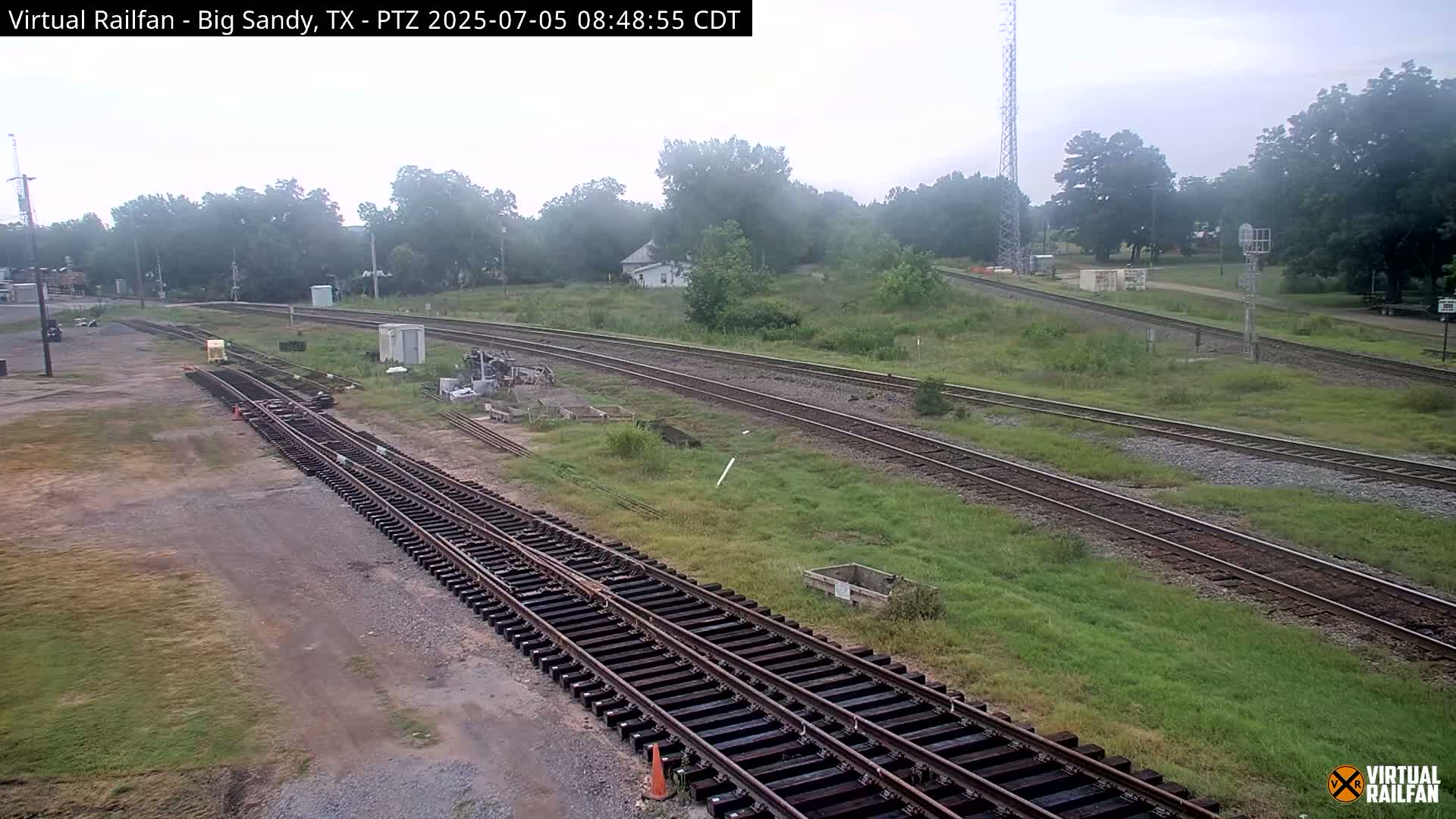 Several sets of train tracks curve through a grassy area with scattered trees and small buildings under an overcast sky.