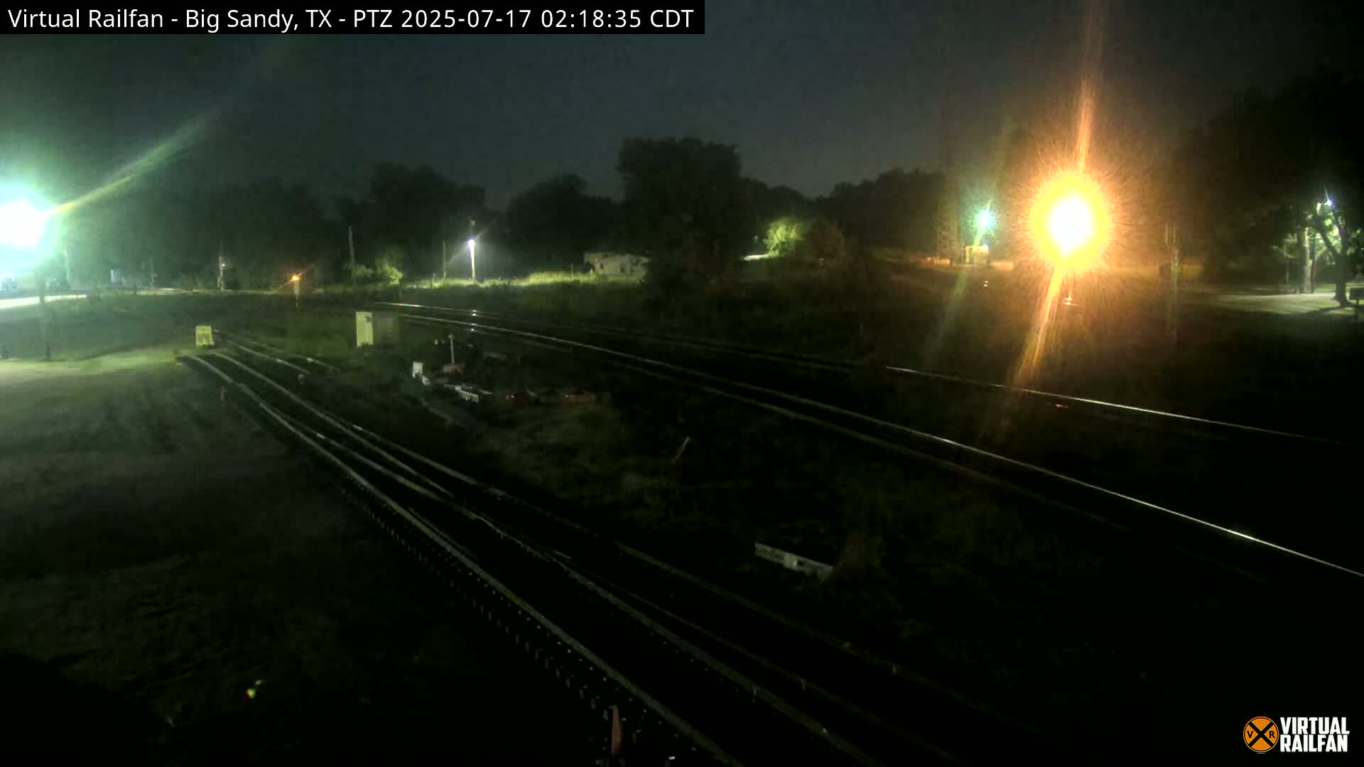 A nighttime view of a railway track system, illuminated by bright lights, surrounded by dark, vegetated areas.