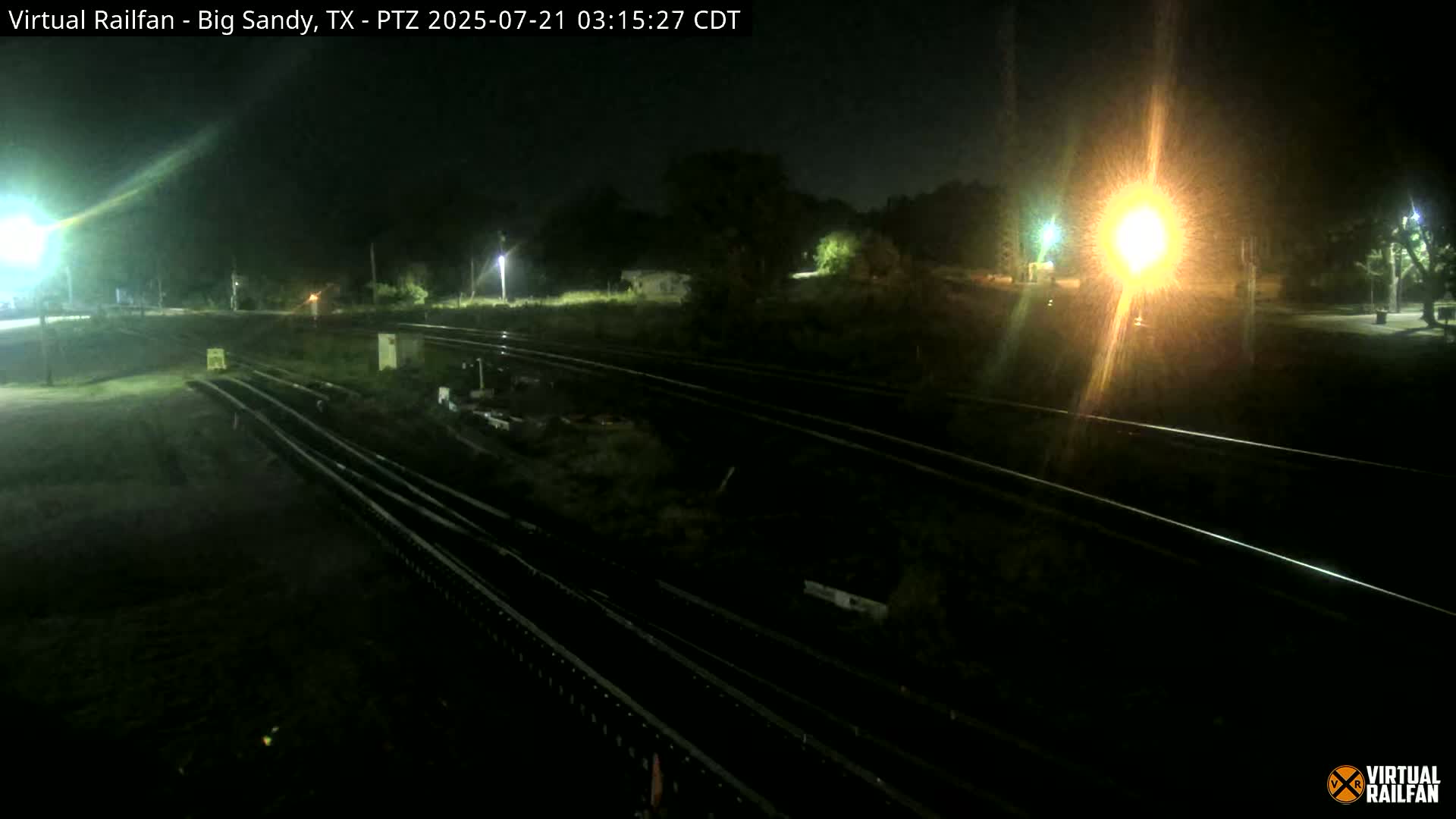 At night, several sets of train tracks are visible, illuminated by bright lights, with some vegetation and structures in the background.