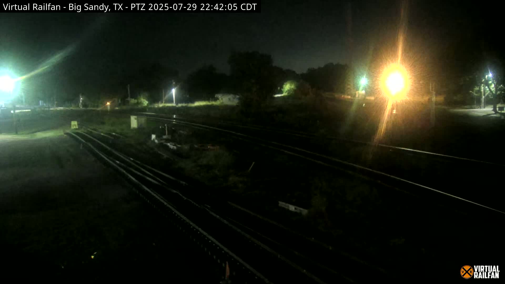 At night, several sets of train tracks are visible, illuminated by bright lights, with some vegetation and structures in the background.