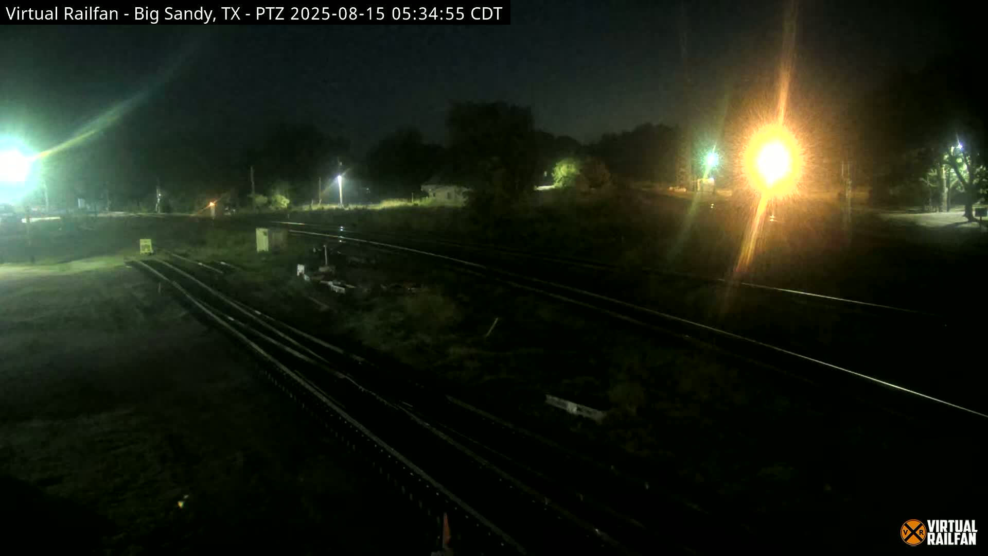 At night, several sets of train tracks are visible, illuminated by bright lights, with some vegetation and structures in the background.