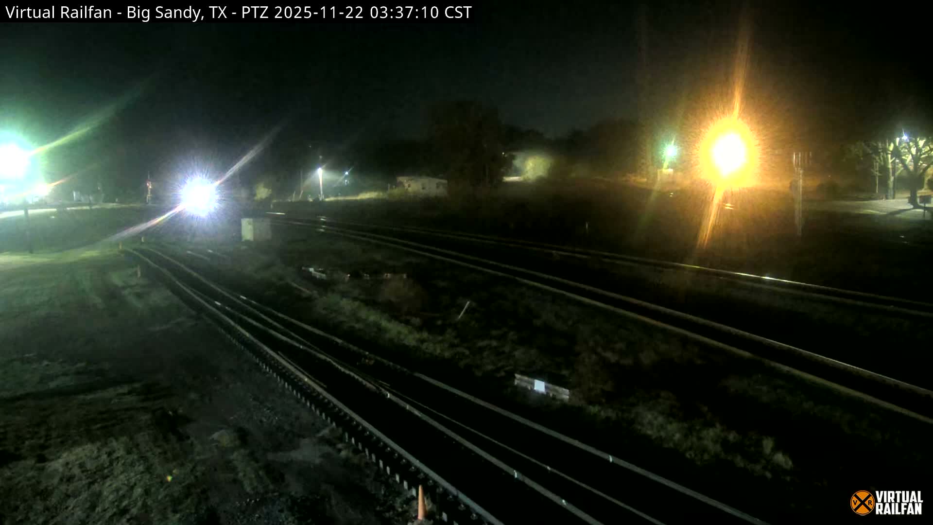 Multiple train tracks stretch across a dark landscape at night, brightly lit by intense headlights on the left and a large, glowing light source on the right, under clear skies.