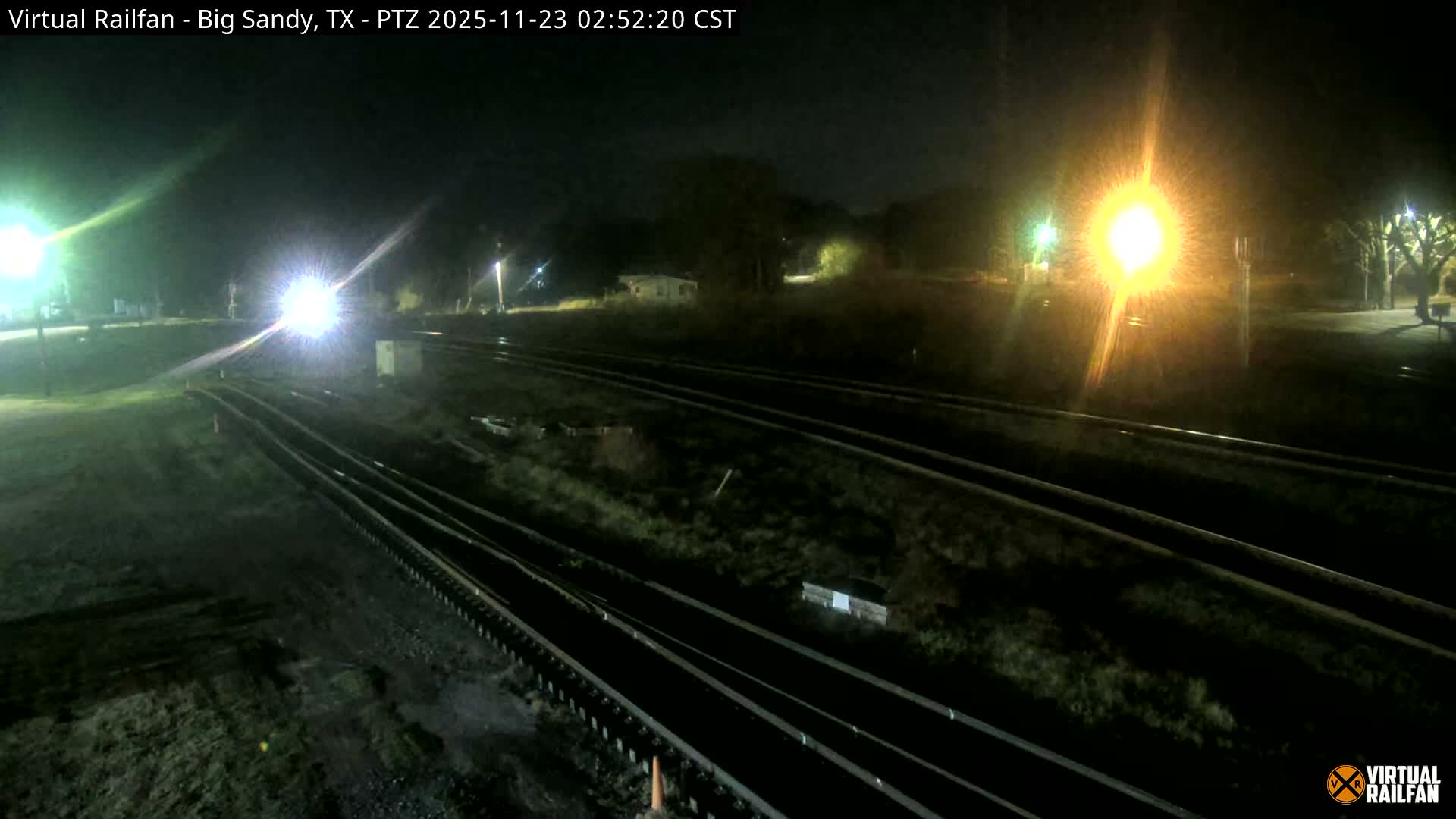 Multiple railroad tracks stretch through an outdoor scene at night, illuminated by several bright lights, including a powerful headlight on the left casting a beam down the tracks.