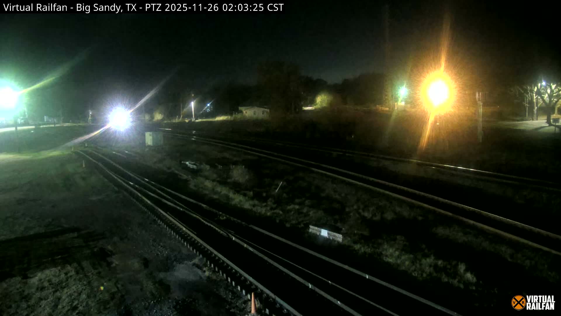 The image shows a dark outdoor night scene with multiple train tracks illuminated by bright artificial lights, including a very bright white light likely from a train's headlights on the left and a prominent yellow light on the right, under clear weather conditions.