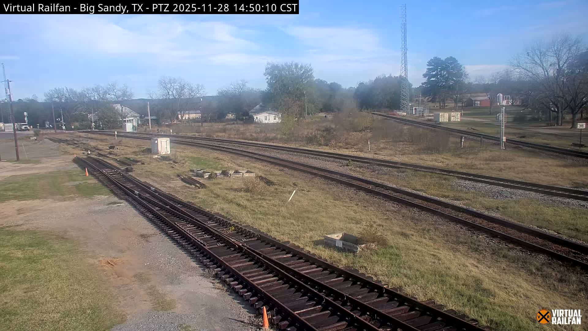 Multiple railway tracks cut through a grassy, semi-rural landscape with scattered bare and evergreen trees, distant buildings, and a communication tower, all beneath a bright, partly cloudy sky.
