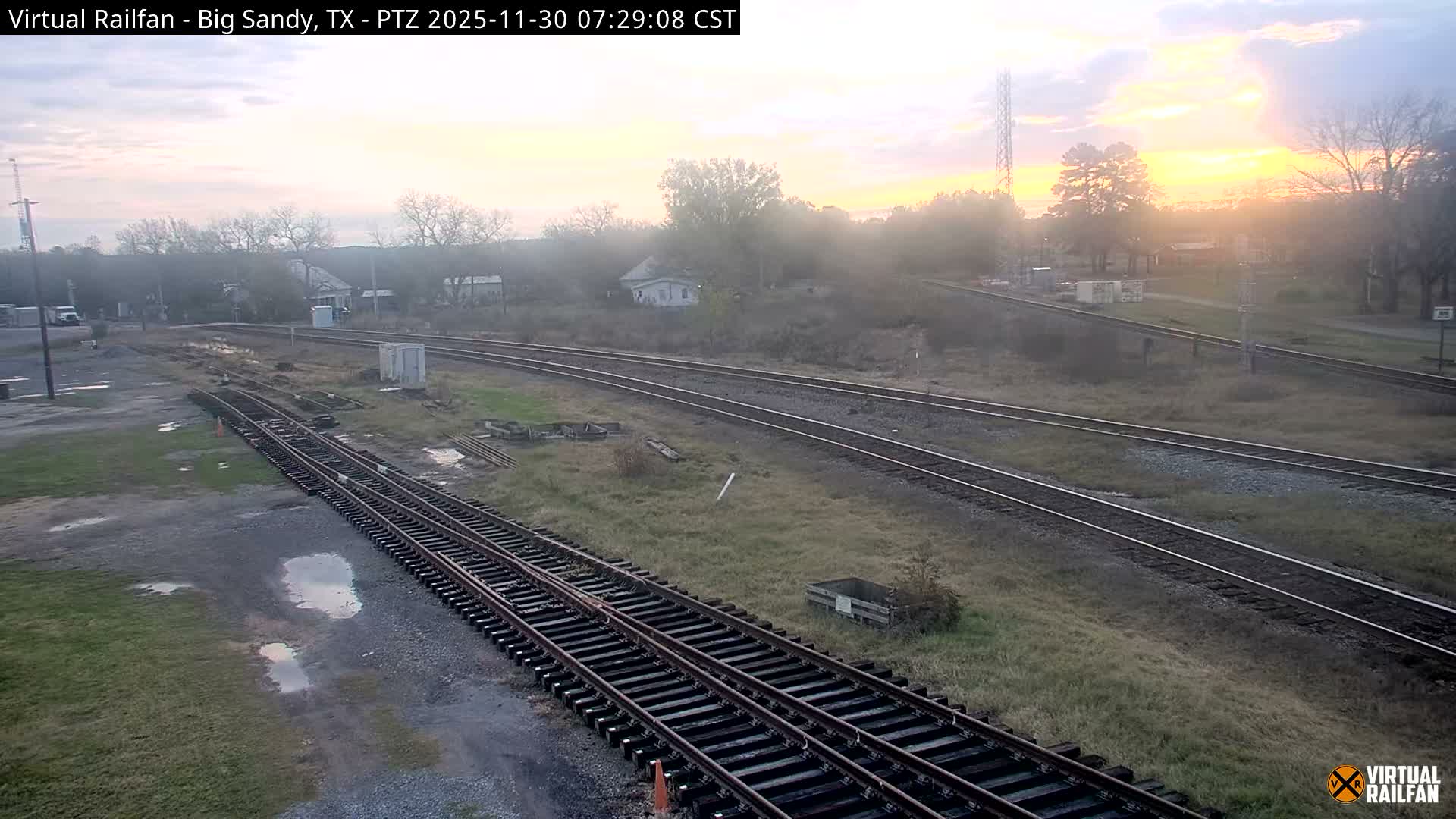 Multiple railway tracks cut through a grassy, semi-rural landscape with scattered bare and evergreen trees, distant buildings, and a communication tower, all beneath a bright, partly cloudy sky.