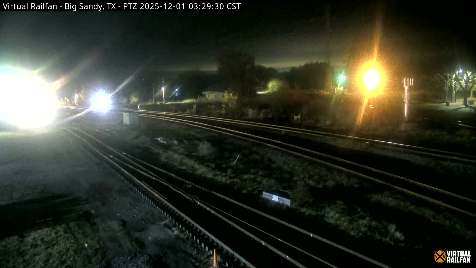 On a clear night, the bright headlights of an approaching train illuminate multiple parallel tracks, with a prominent orange signal light glowing on the right.