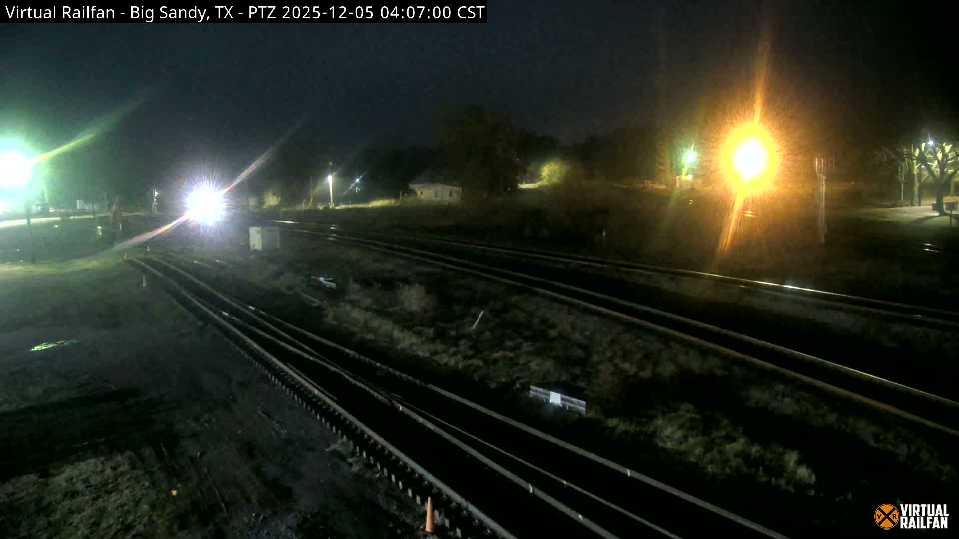 Multiple train tracks converge under a dark, clear night sky, illuminated by several bright lights, one prominently resembling a train headlight.