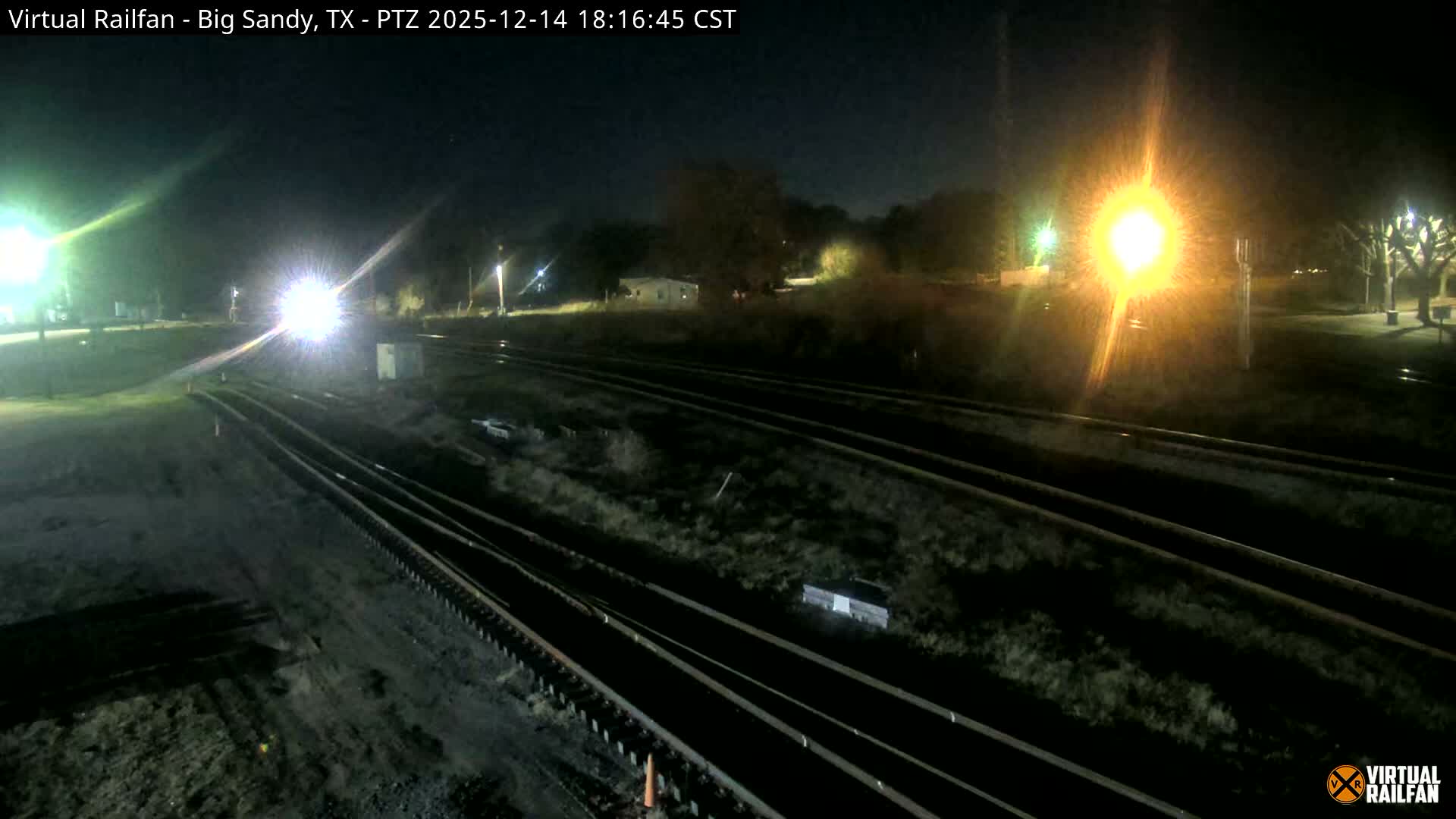 Multiple train tracks converge under a dark, clear night sky, illuminated by several bright lights, one prominently resembling a train headlight.