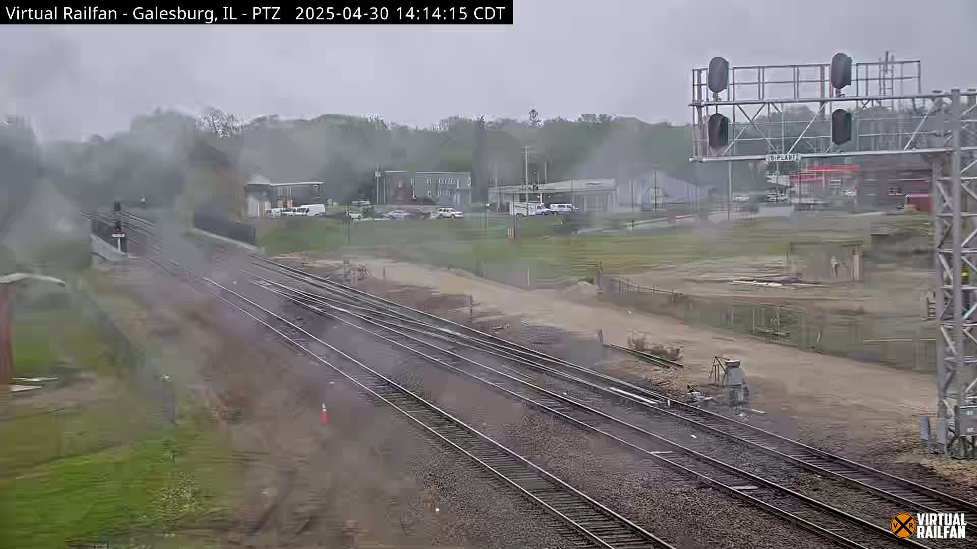 A steam train is emitting smoke as it passes through a railroad yard on an overcast day.