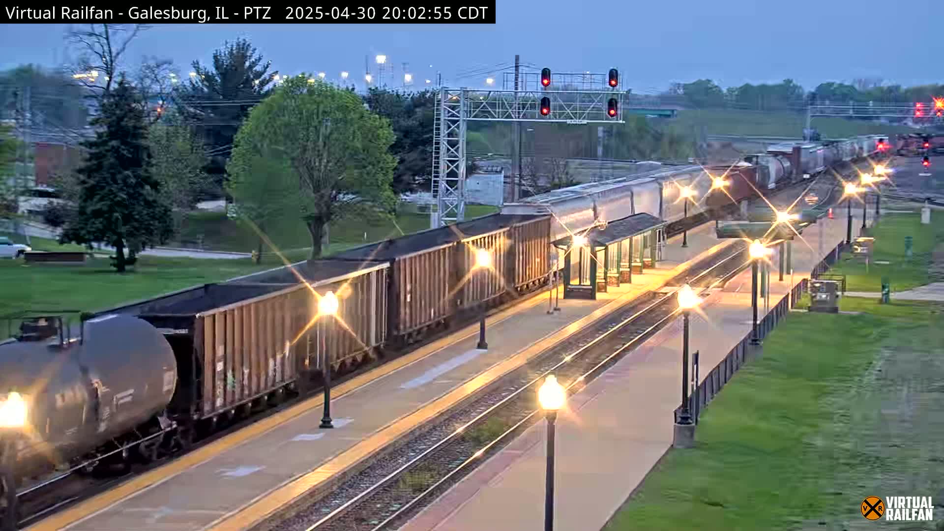 A long freight train passes a train station at dusk, under a twilight sky.