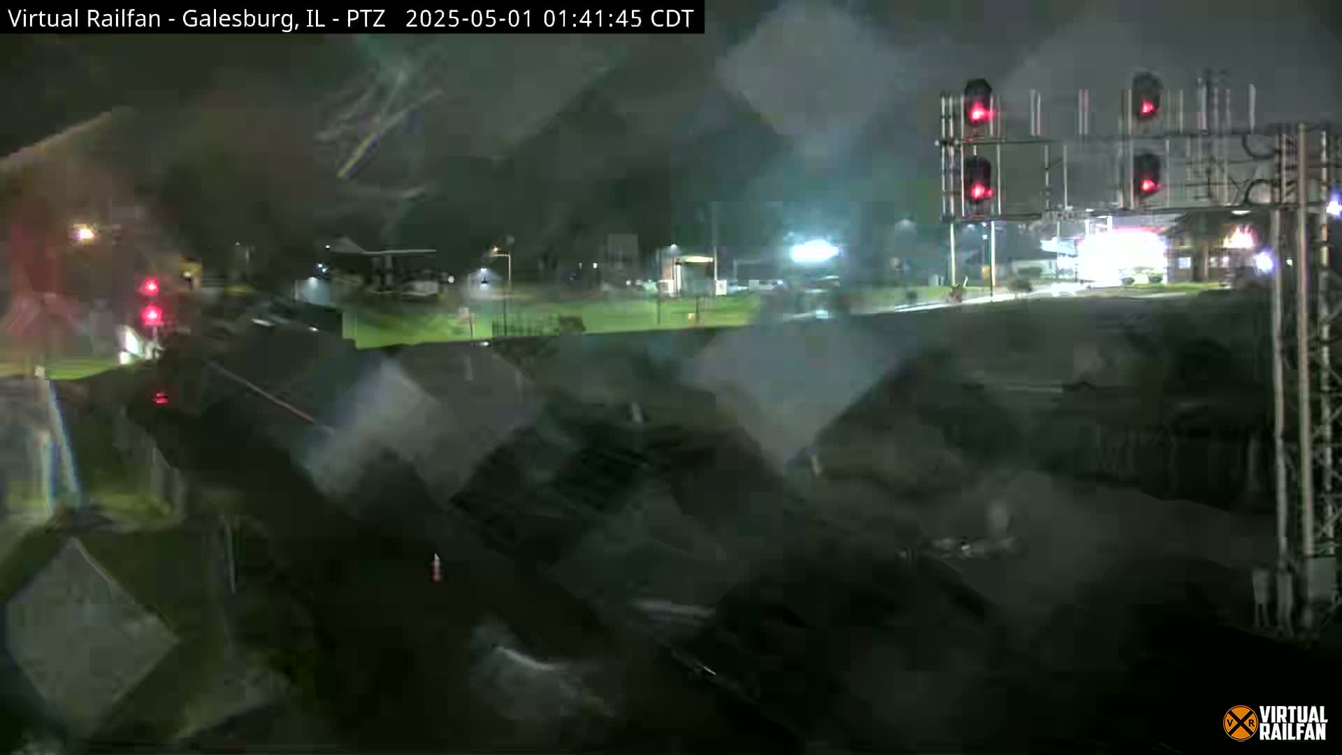 A nighttime view of a rain-slicked road and railroad crossing with a train partially visible, illuminated by streetlights and the red lights of a railroad crossing signal.