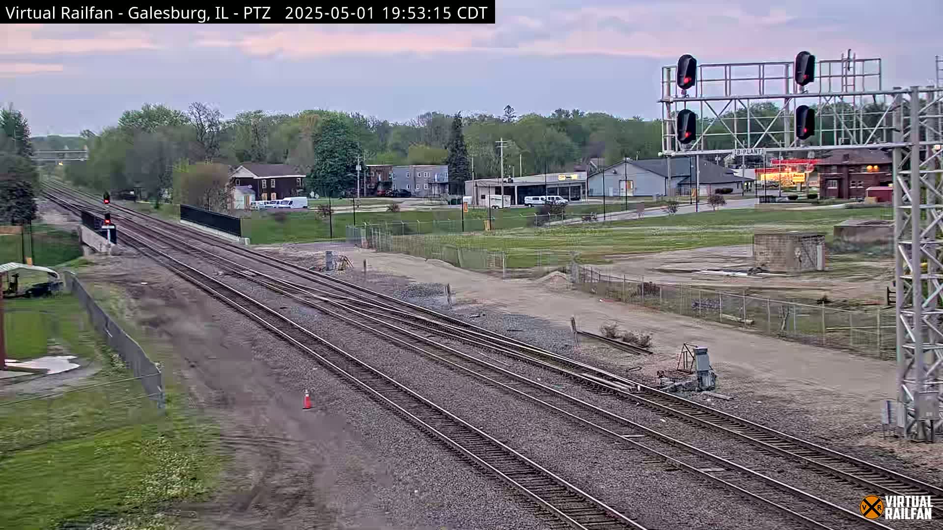 Multiple sets of train tracks extend into the distance under a twilight sky, passing through a flat landscape with some buildings and trees visible in the background.