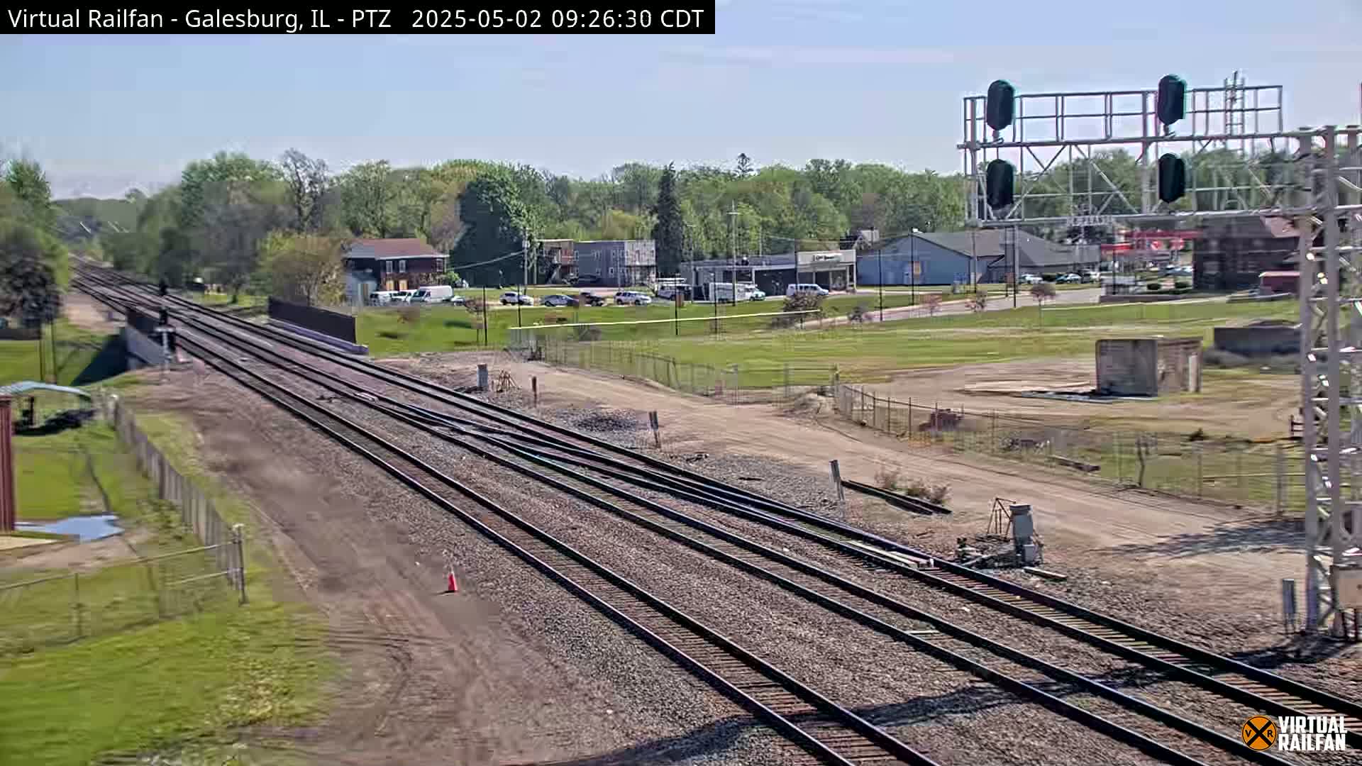 Multiple train tracks stretch into the distance under a bright, sunny sky, passing in front of a small town with low buildings and green spaces.