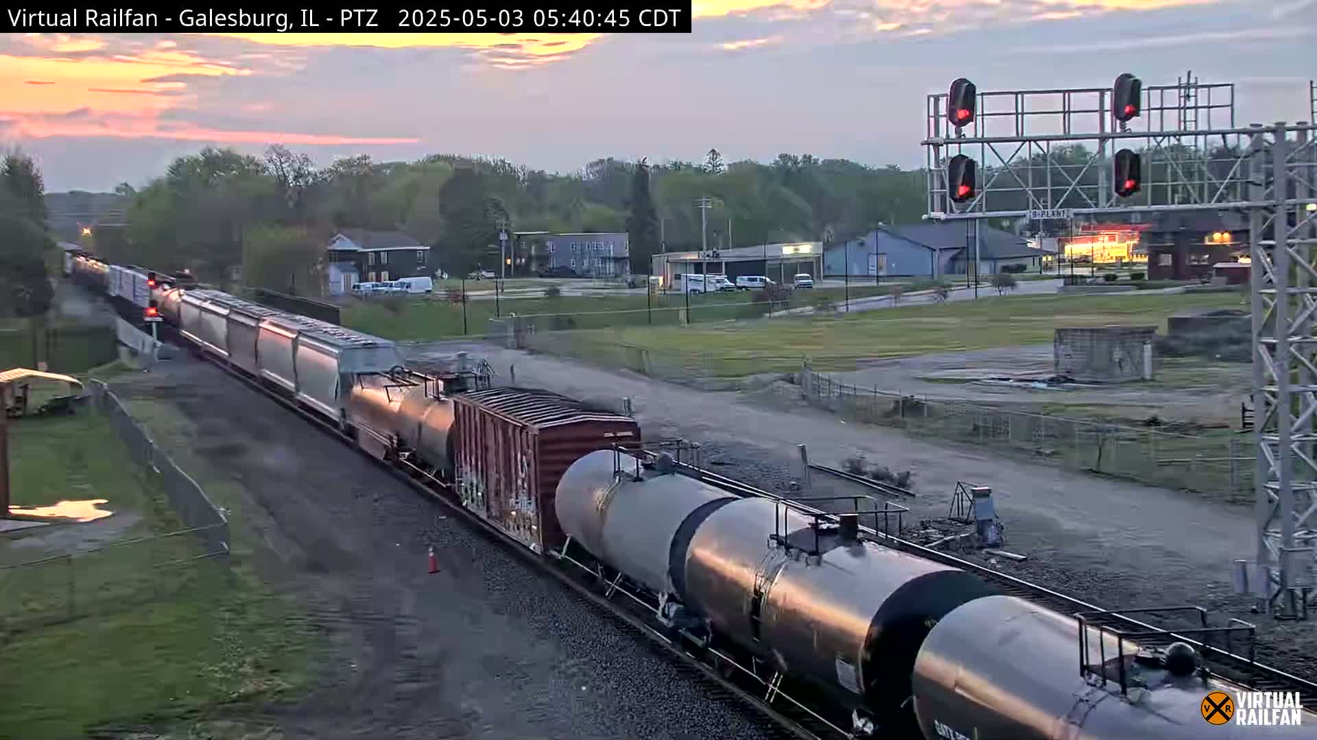 A long freight train passes under a railway signal during a sunrise with a partly cloudy sky.