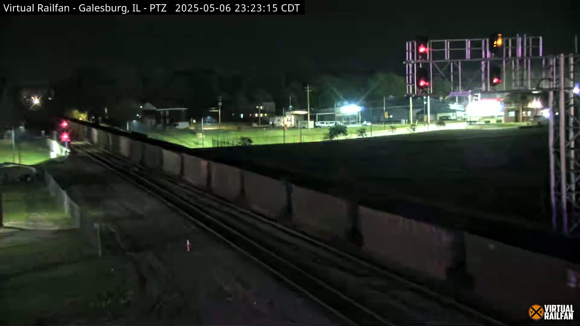 A long freight train passes under a set of railway signals at night.