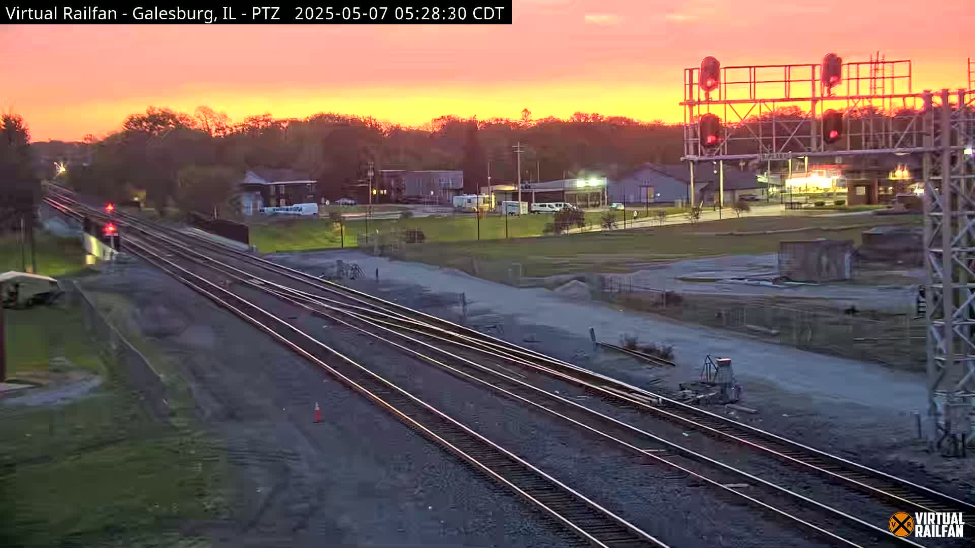 Multiple sets of train tracks extend into the distance under a vibrant orange and pink sunrise, passing through a flat landscape with some buildings and sparse vegetation.