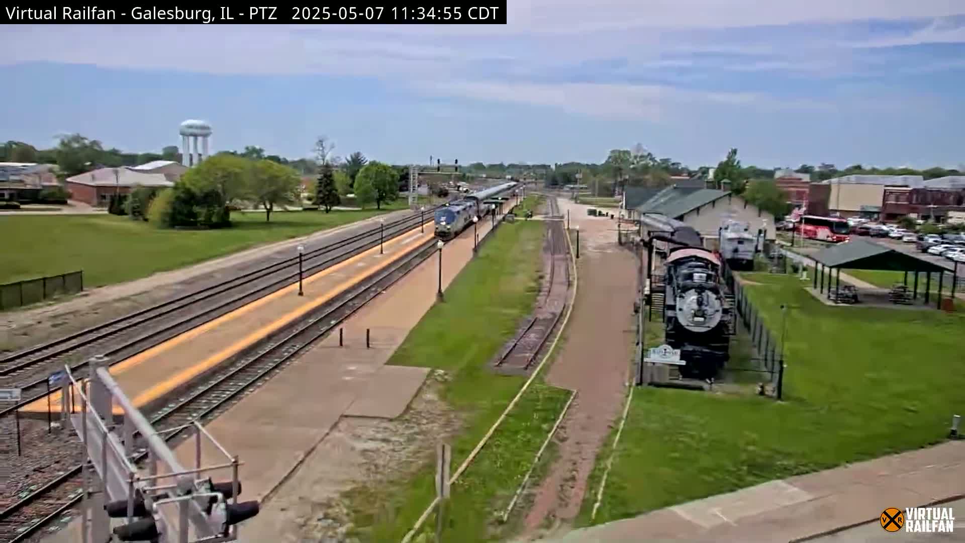 A passenger train is passing through a train station on a partly sunny day, with a vintage locomotive displayed nearby.
