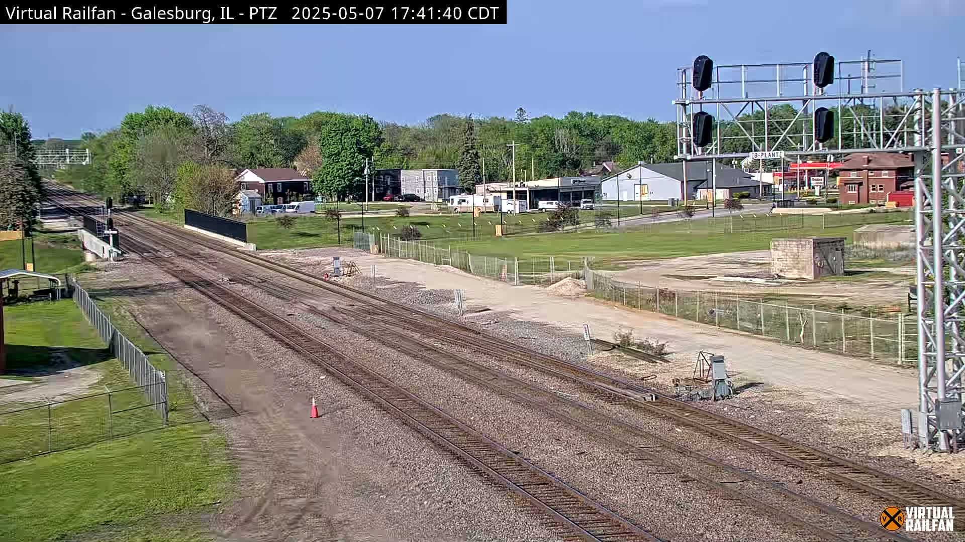 Multiple sets of train tracks stretch into the distance under a clear blue sky, passing through a town with low buildings and green spaces.