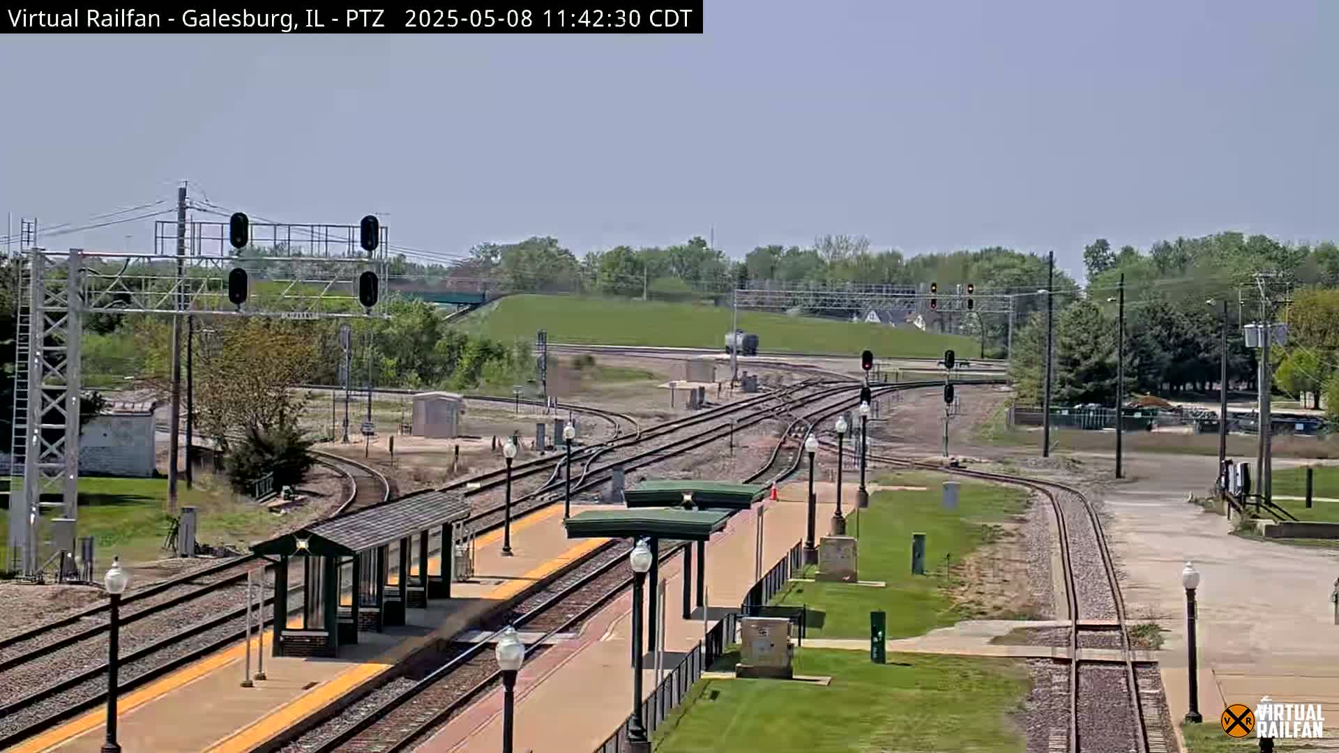 A train station with multiple train tracks and a covered waiting area is seen on a sunny day.