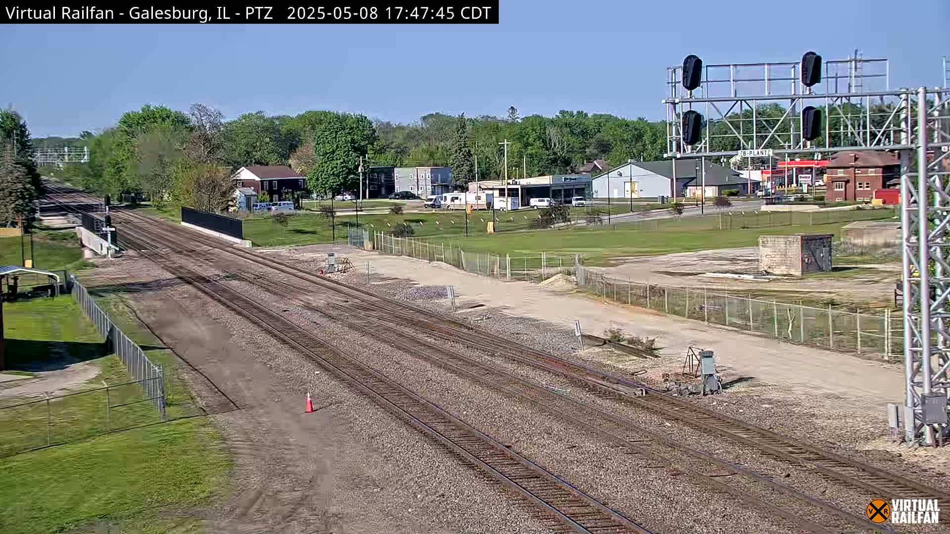 Multiple train tracks stretch into the distance under a clear blue sky, passing through a suburban area with low buildings and green grass.