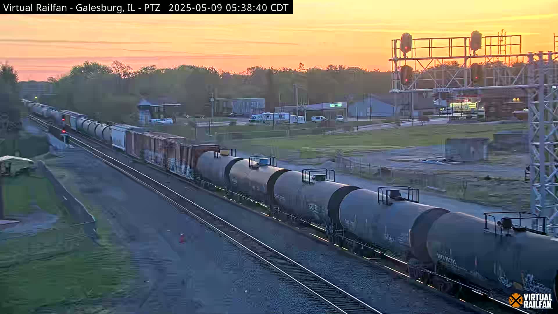 A long freight train carrying tanker cars passes under a railroad signal at sunrise.