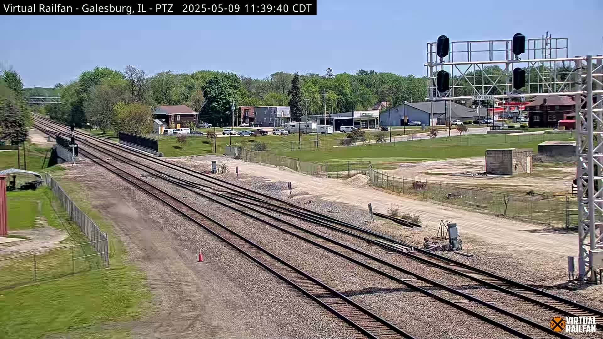 Multiple train tracks stretch into the distance on a sunny day, past a small town with buildings and sparse vegetation.