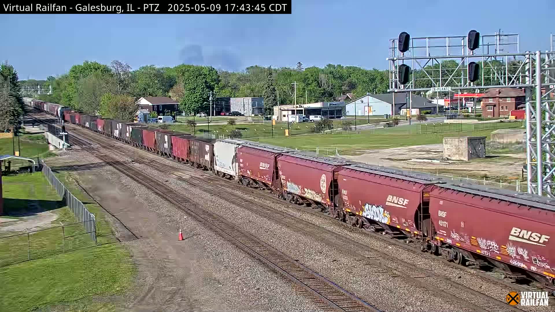 A long freight train of red and black cars travels along railroad tracks on a sunny day, passing through a suburban area with trees and buildings in the background.