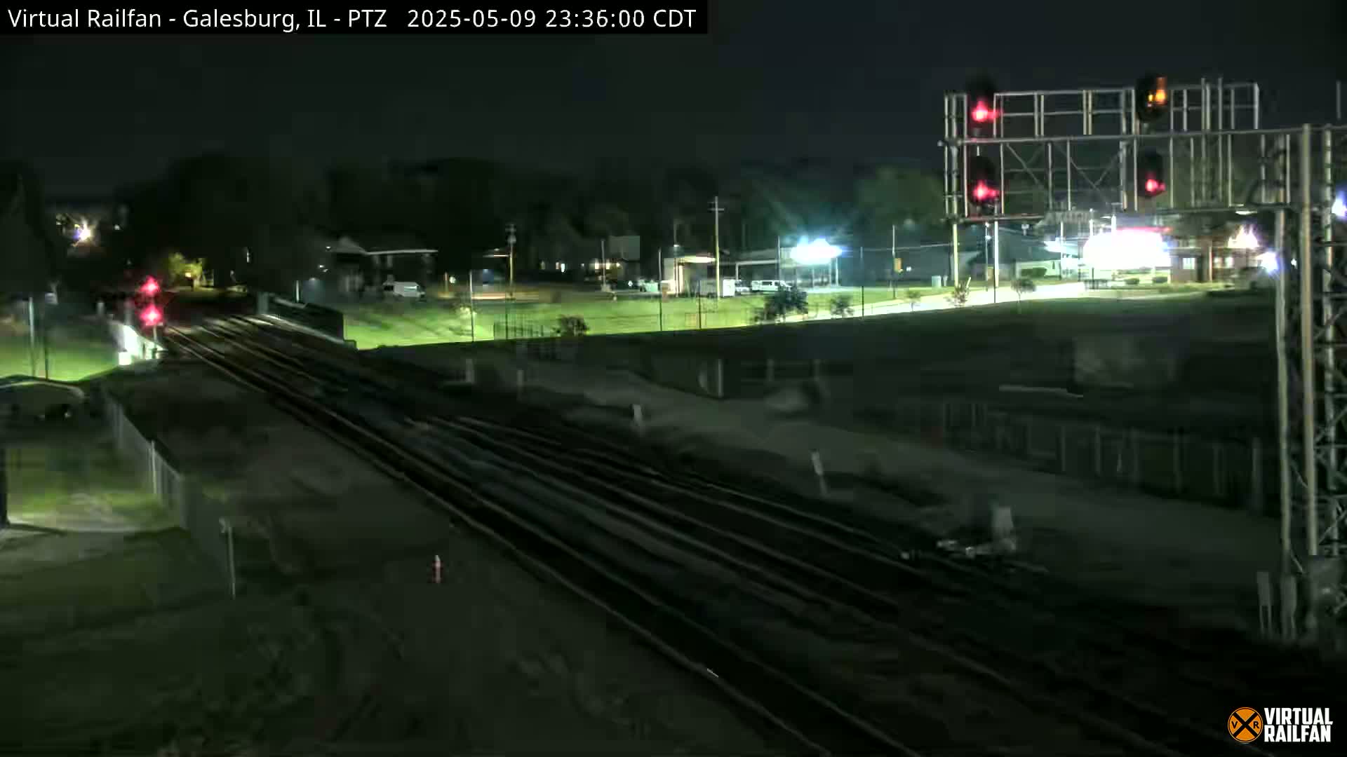 A nighttime view of several train tracks crossing under a road, with red traffic signals illuminated and minimal surrounding traffic.