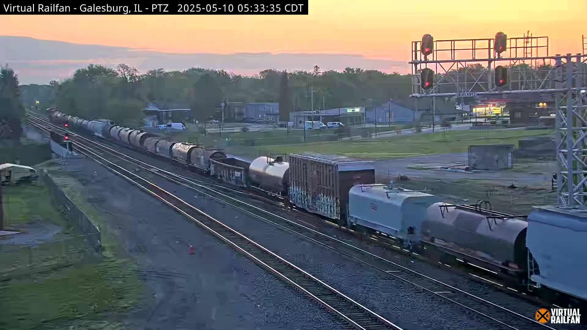 A long freight train passes through a flat landscape at sunrise under a pale orange sky.