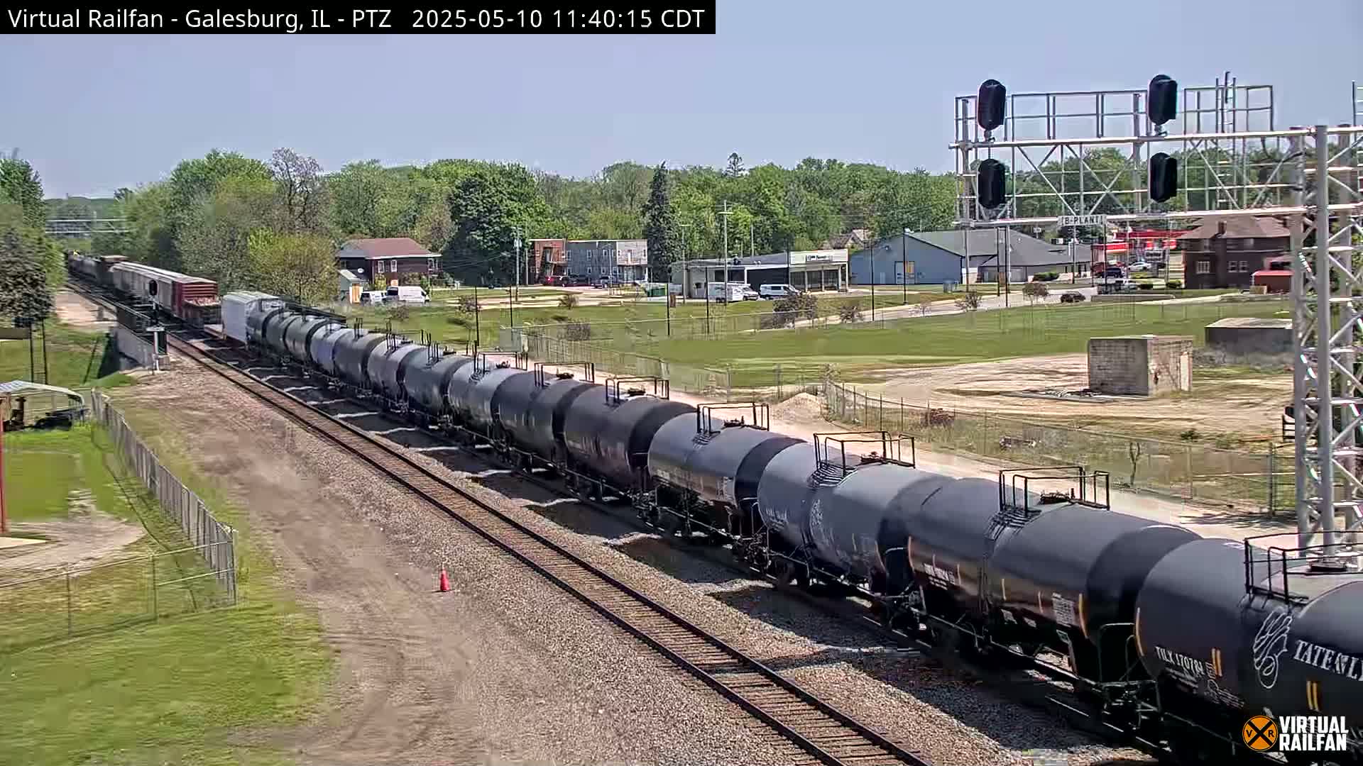 A long freight train of tank cars travels along railroad tracks on a sunny day past a small town.