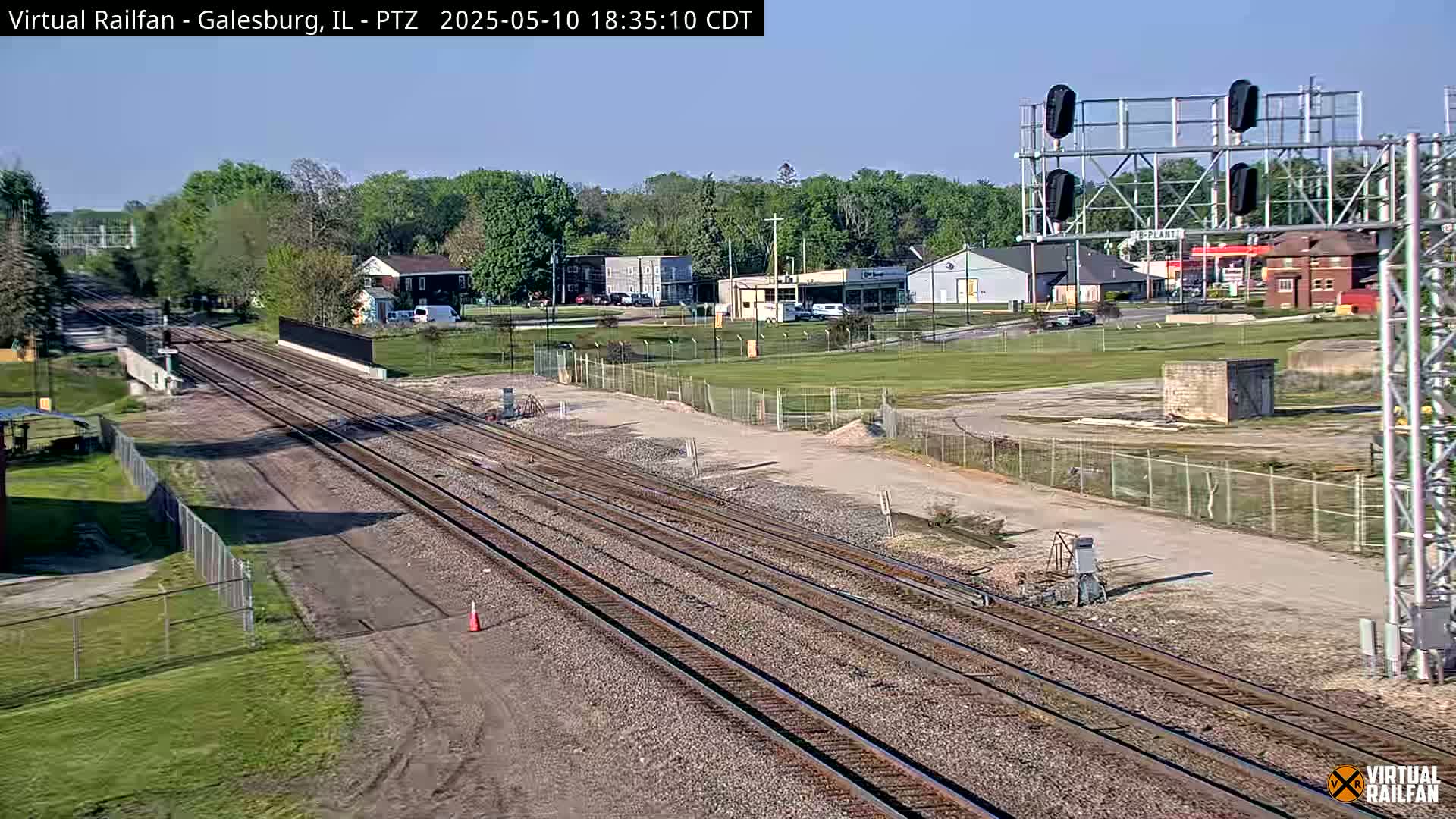 Multiple train tracks extend into the distance under a clear blue sky, passing through a grassy area with buildings and trees in the background.