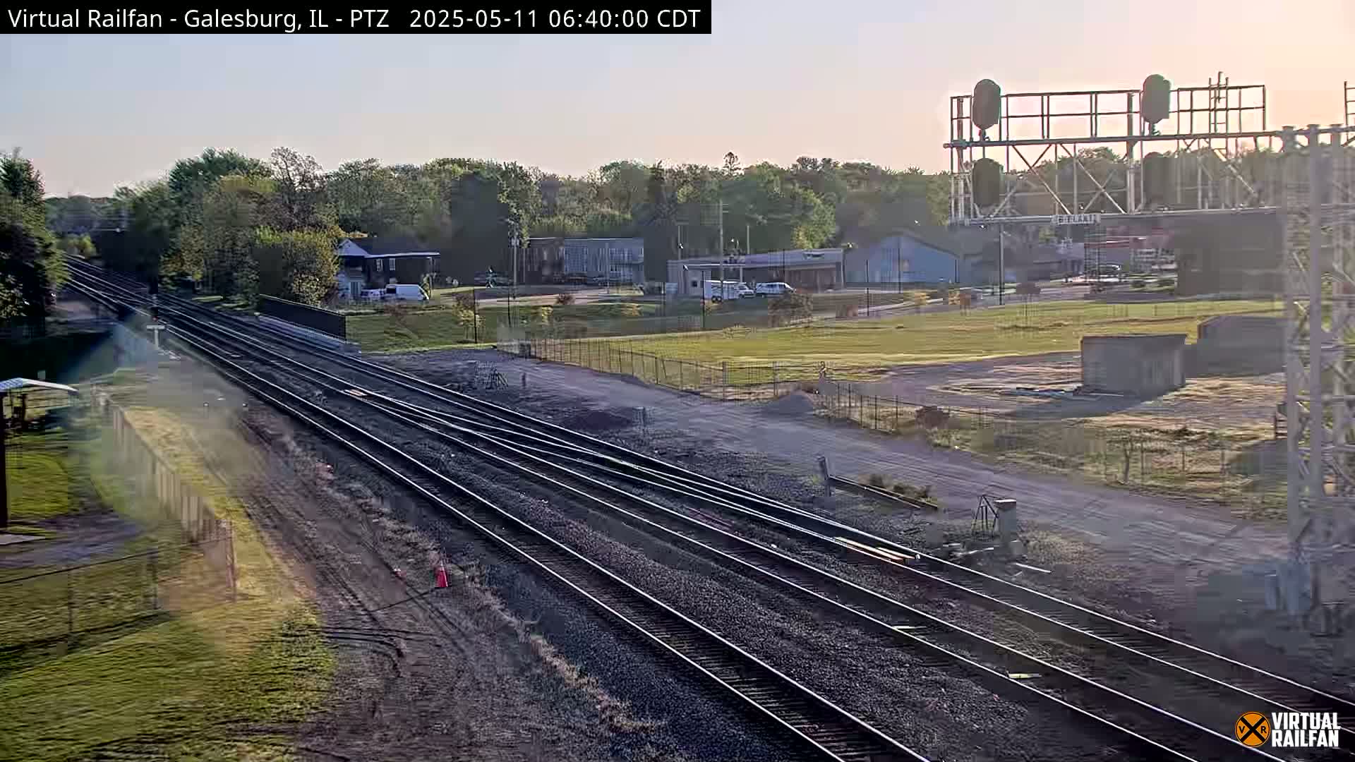 Multiple train tracks stretch into the distance under a clear, bright sky, passing by a grassy field and some low buildings.