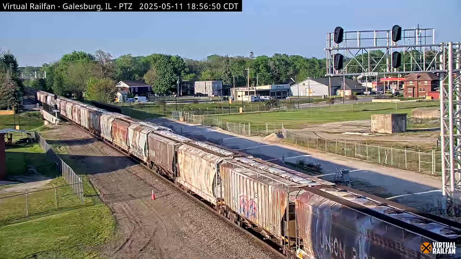 A long freight train with graffiti-covered cars moves along tracks past a grassy area and buildings under a clear, sunny sky.