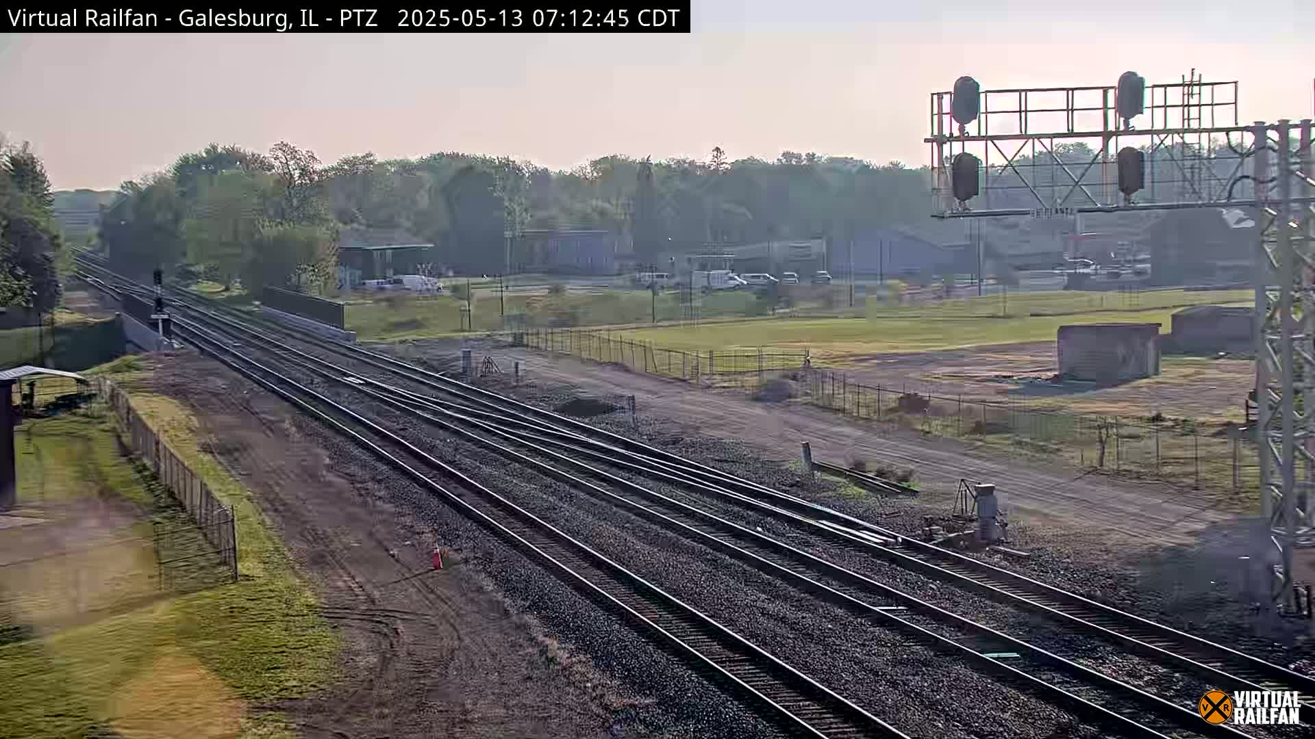 Multiple train tracks extend into the distance under a hazy, sunny sky, past a grassy field and some industrial buildings.
