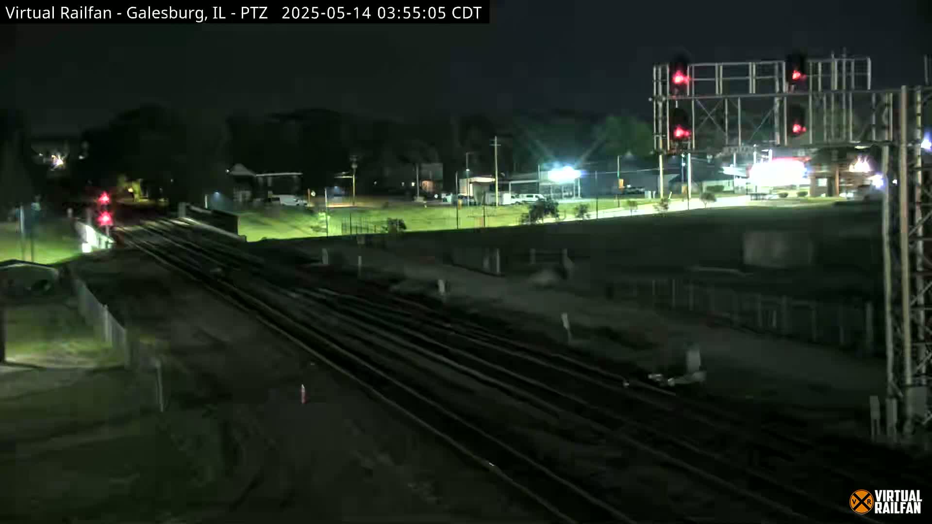A nighttime view of railroad tracks and a signal, with several buildings visible in the background under a clear sky.