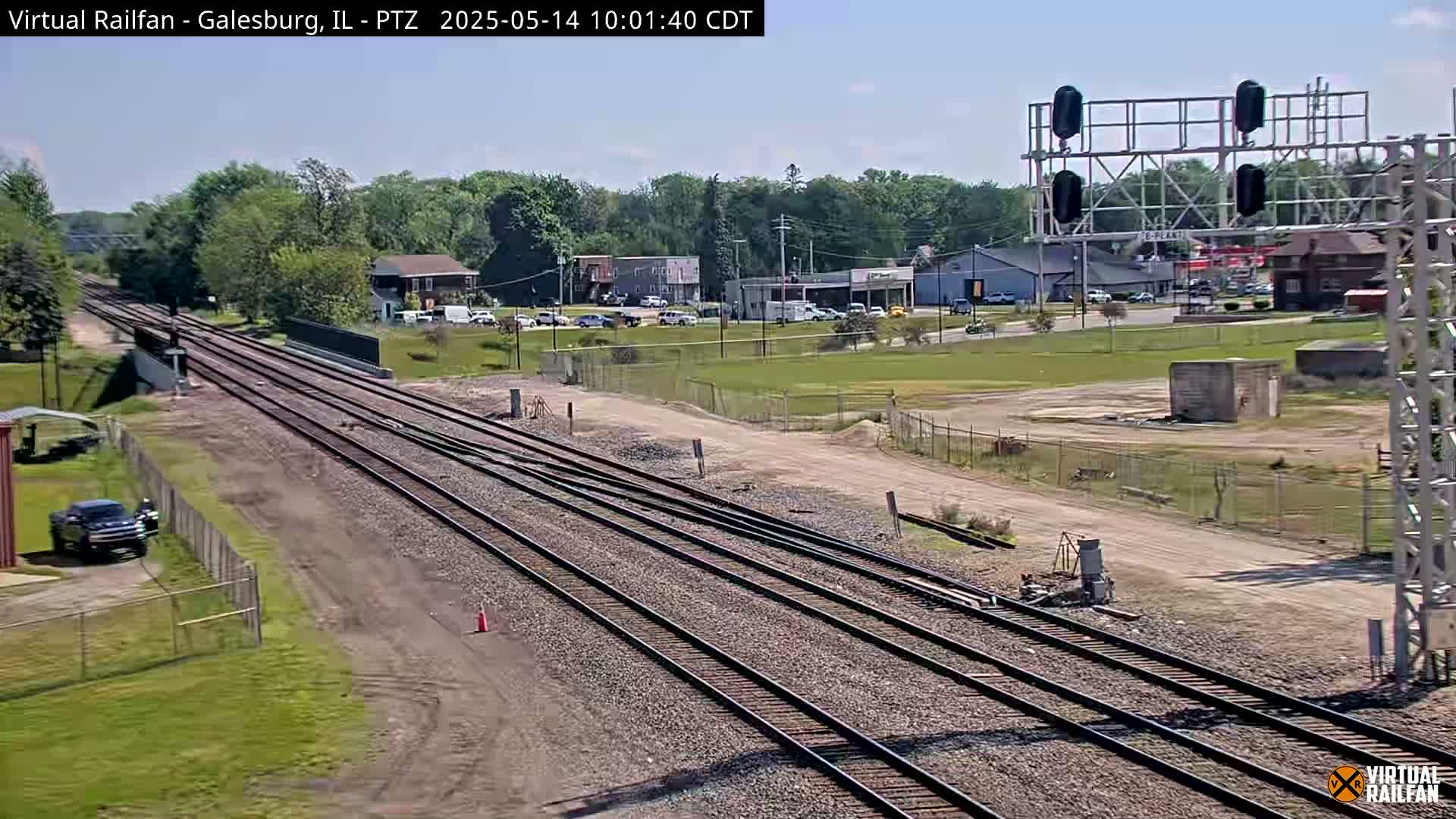 Multiple train tracks stretch into the distance under a sunny sky, past a roadway with buildings and cars visible in the background.
