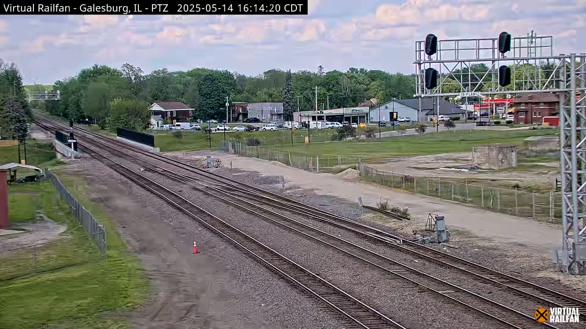 Multiple train tracks extend into the distance under a partly cloudy sky, passing by a small town with buildings and parked cars.