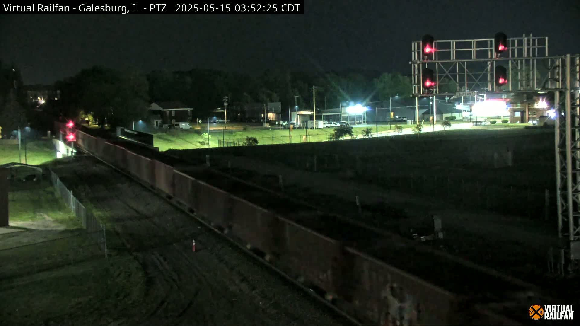 A long freight train passes under a set of red railroad crossing lights at night.