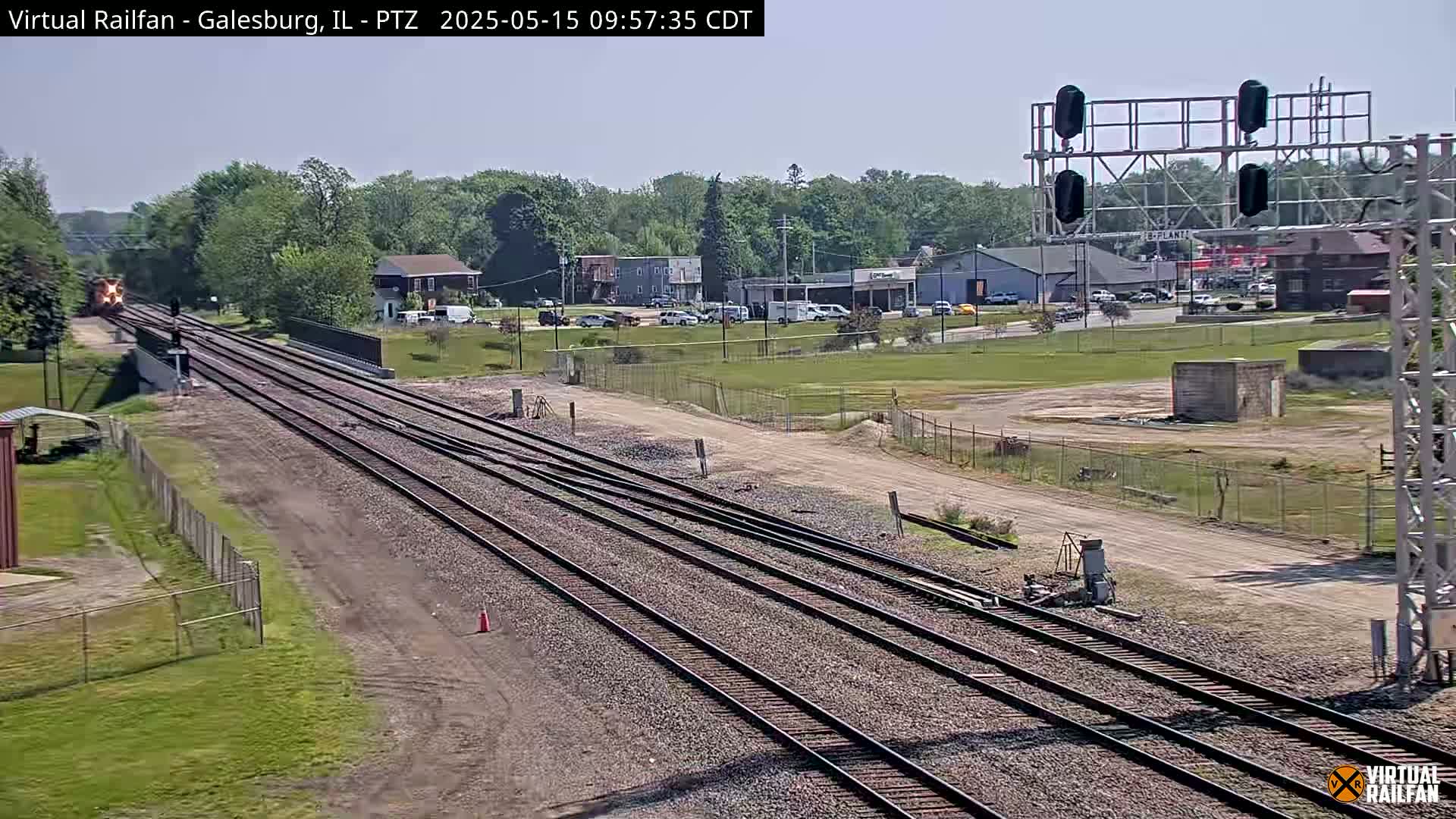 A train passes under a railroad crossing signal on a sunny day, with multiple sets of tracks extending into the distance past a grassy area and buildings.