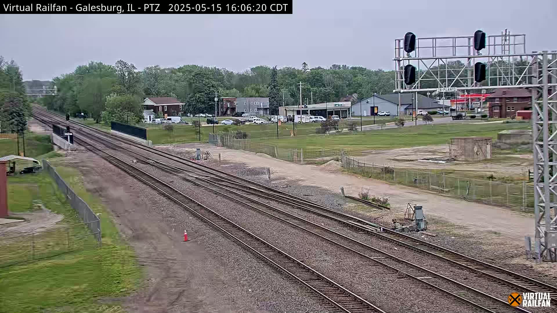 Multiple sets of train tracks stretch into the distance under an overcast sky, passing by a road with buildings and cars visible in the background.