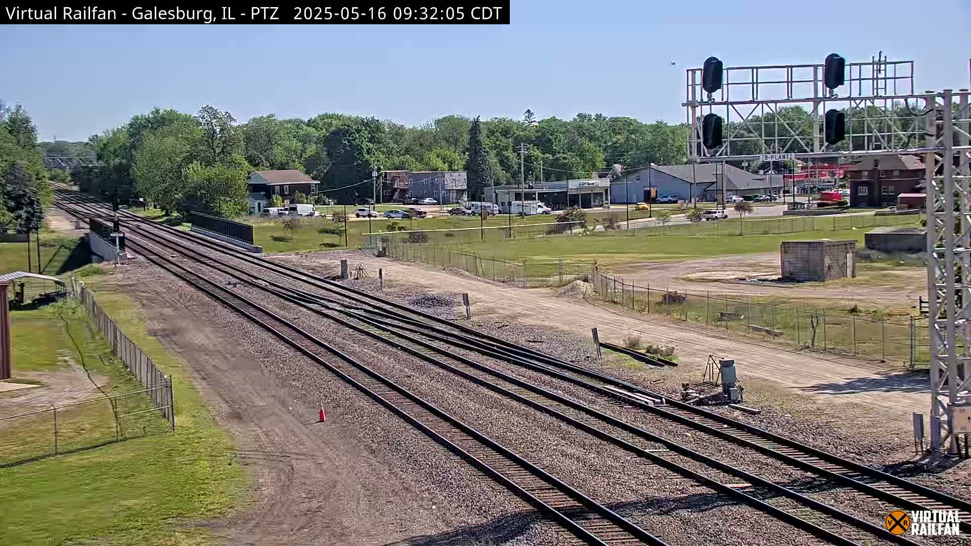Multiple sets of train tracks extend into the distance under a clear sky, past a roadway and buildings.