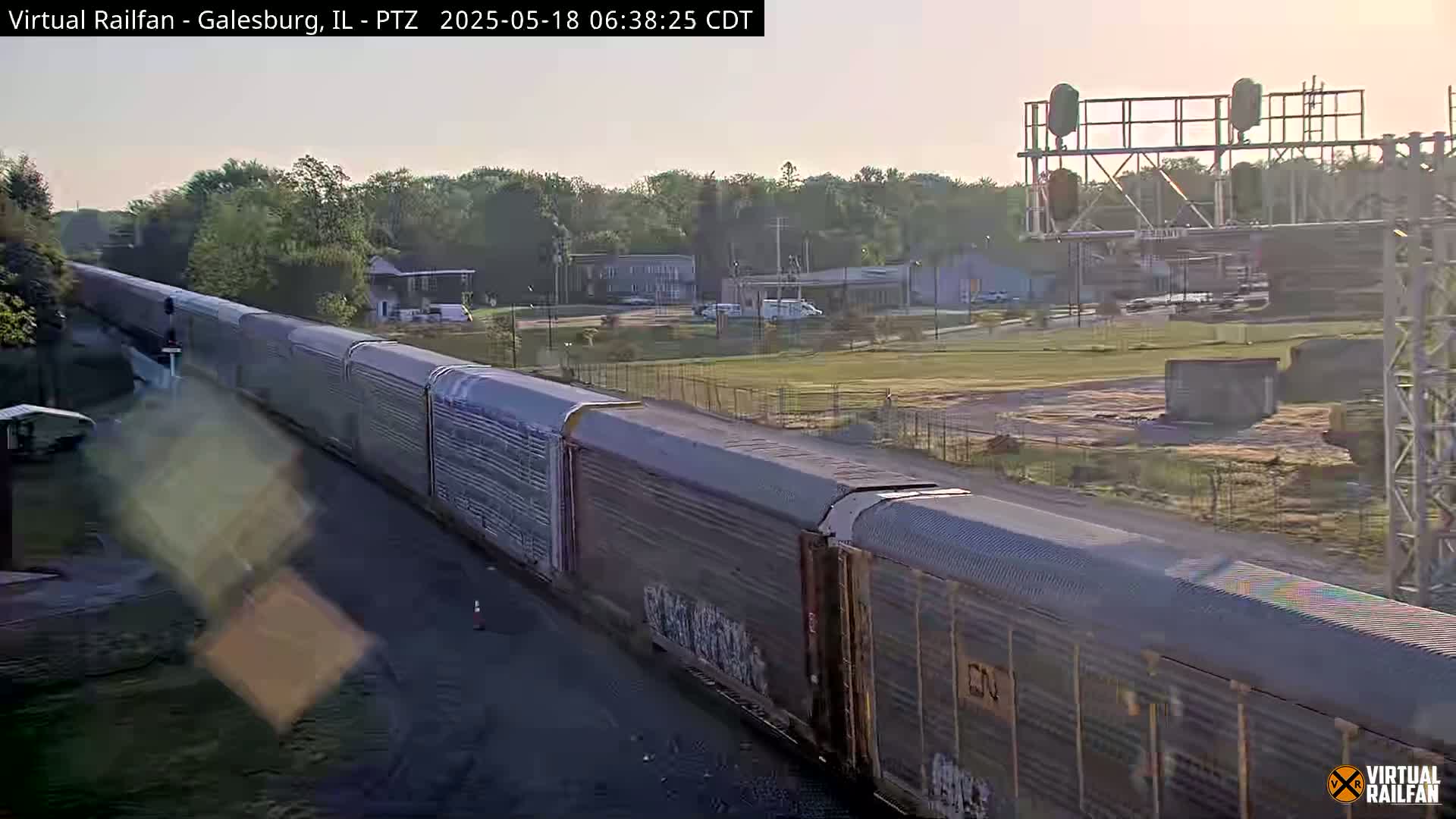 A long freight train passes by under a mostly clear morning sky, with trees and buildings visible in the background.