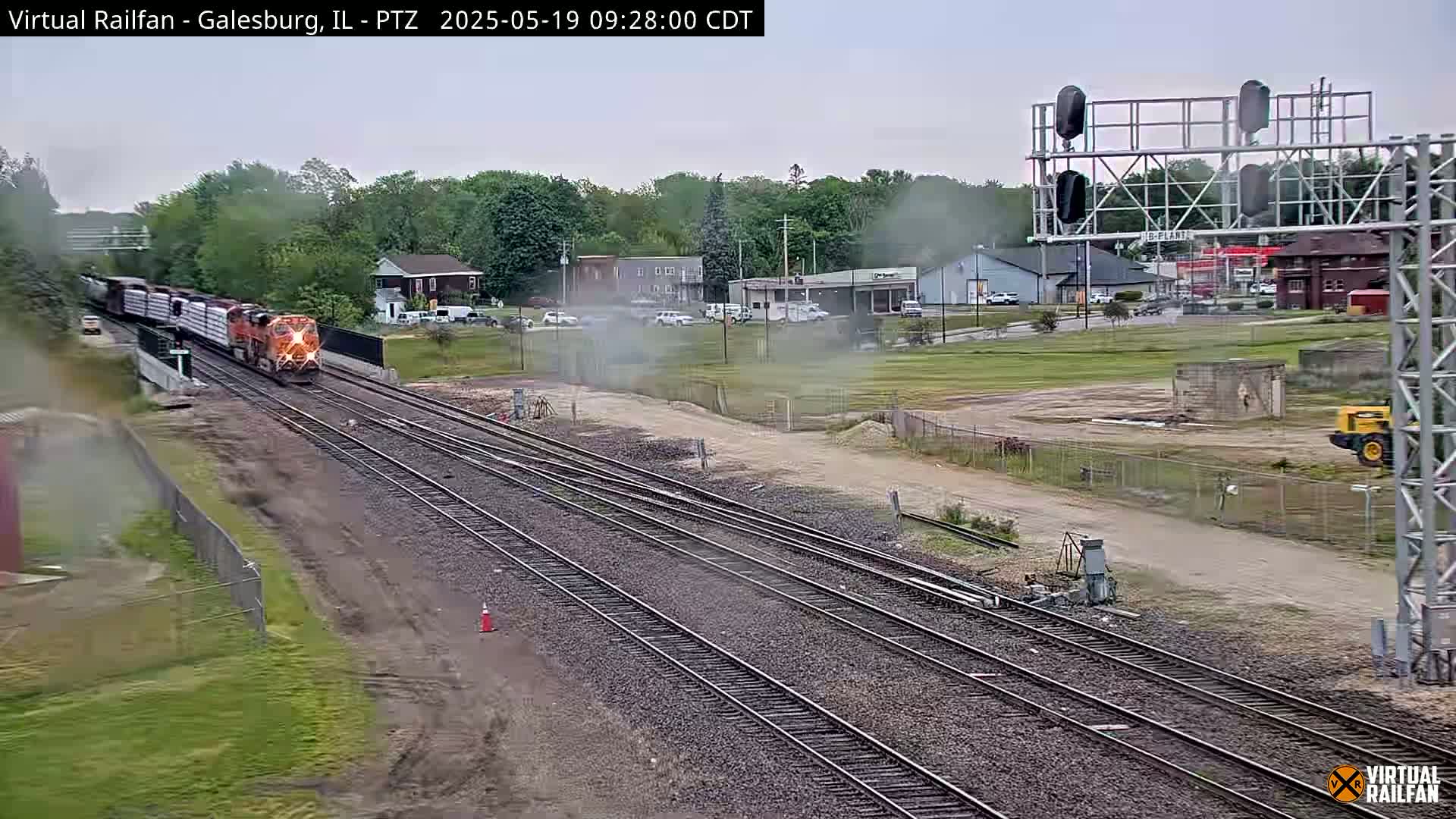A long freight train passes by a railway junction on an overcast day, near a small town with some light construction visible.